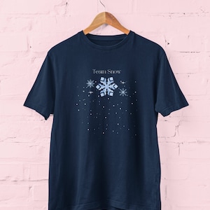 May include: A navy blue t-shirt with the words "Team Snow" and a snowflake design. The design includes a large, light blue snowflake, smaller snowflakes, and white dots. The t-shirt is displayed on a wooden hanger against a pink brick wall.