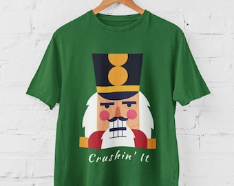 Può includere: T-shirt verde con un disegno di schiaccianoci a cartoni animati. Lo schiaccianoci ha un cappello nero con dettagli dorati, barba bianca e cappotto rosso. Il testo "Crushin' It" è stampato sotto.