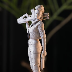 Può includere: Una statuetta bianca stampata in 3D di una donna con un taglio di capelli mohawk. Indossa una giacca, pantaloni e stivali. Tiene una pistola in una mano e un fucile nell'altra.