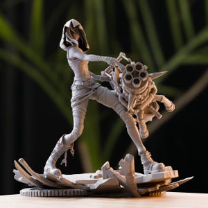 May include: A grey figurine of a character in a dynamic pose, holding a large weapon. The figure is wearing a hooded top, wrapped bandages, and boots. The base is a complex, fragmented design.