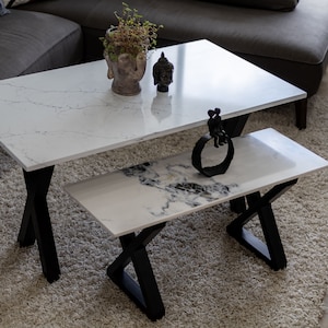 May include: A white marble coffee table with black X-shaped legs and a smaller white marble bench with black X-shaped legs. The table has a white marble top and a black metal sculpture of two people sitting on a bench.