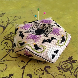 May include: A beige fabric pincushion with a black spiderweb design, purple bats, and black birds. The pincushion is shaped like a cube and has a black button in the centre. There are four colourful pins stuck in the top of the pincushion.