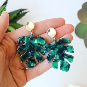 May include: A pair of green and black monstera leaf earrings with gold accents. The earrings are made of resin and have a marbled effect.