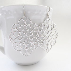 Puede incluir: Un par de pendientes de filigrana plateada con un dise&ntilde;o delicado e intrincado. Los pendientes cuelgan de una taza blanca.