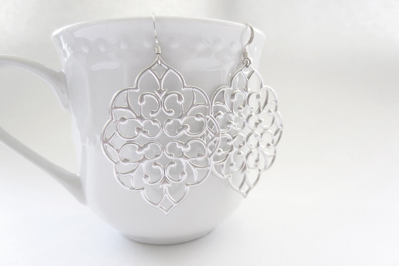 Puede incluir: Un par de pendientes de filigrana plateada con un dise&ntilde;o delicado e intrincado. Los pendientes cuelgan de una taza blanca.