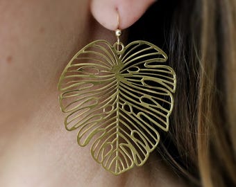 May include: Gold-tone earrings featuring a large, detailed cutout of a monstera leaf.