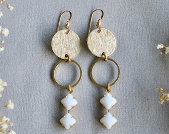 May include: Gold earrings with two white square-shaped stones hanging from a gold circle. The top of the earrings feature a gold circle with a textured surface.