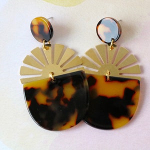 Pode incluir: Um par de brincos de ouro e tartaruga com um design de sol. Os brincos s&atilde;o feitos de metal e resina.