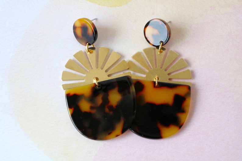 Pode incluir: Um par de brincos de ouro e tartaruga com um design de sol. Os brincos s&atilde;o feitos de metal e resina.