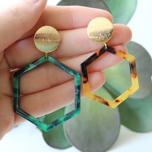 May include: A pair of gold and green geometric earrings. The earrings are hexagonal shaped with a gold disc at the top. The green earring has a marbled pattern.