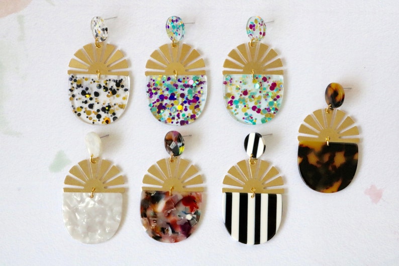 Pode incluir: Seis pares de brincos dourados com um design de sol. Cada brinco apresenta um padr&atilde;o diferente na metade inferior, incluindo listras pretas e brancas, tartaruga, branco marmorizado e glitter de v&aacute;rias cores.