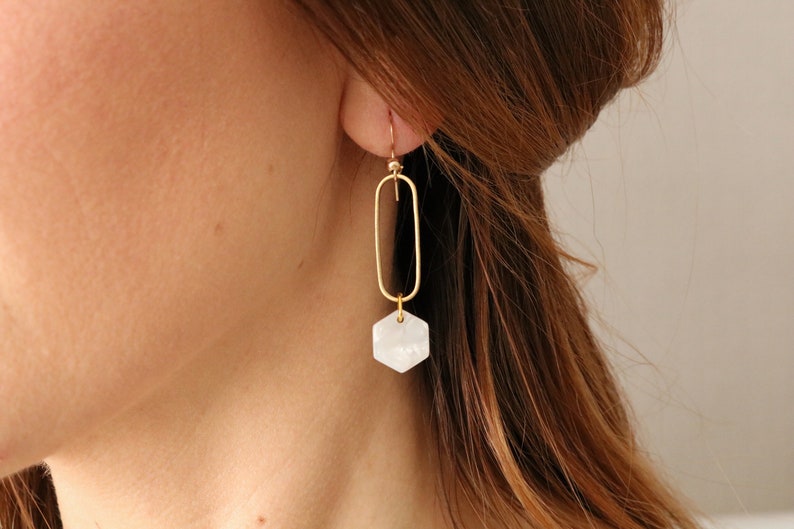 May include: Gold oval hoop earrings with a white hexagonal pendant. The pendant has a textured surface.