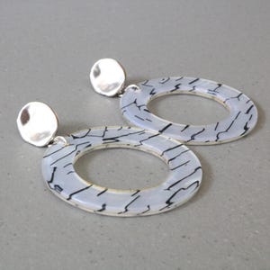 May include: A pair of silver and white hoop earrings with a black marbled design. The earrings are large and have a circular shape.