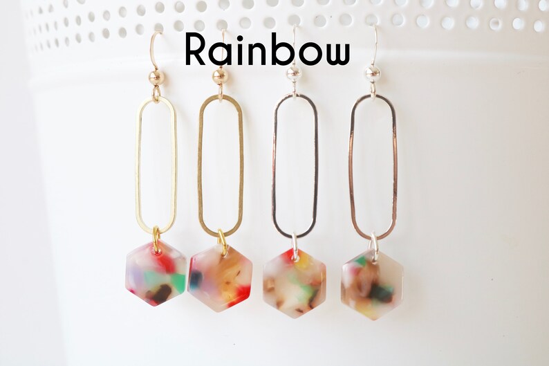 May include: Four pairs of dangle earrings with a hexagonal charm. The charms are made of clear resin with colourful flecks. The earrings are available in gold, silver, and rose gold.
