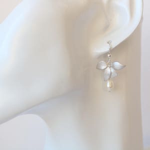 May include: Silver flower earrings with a single pearl drop. The earrings are on a white mannequin head.