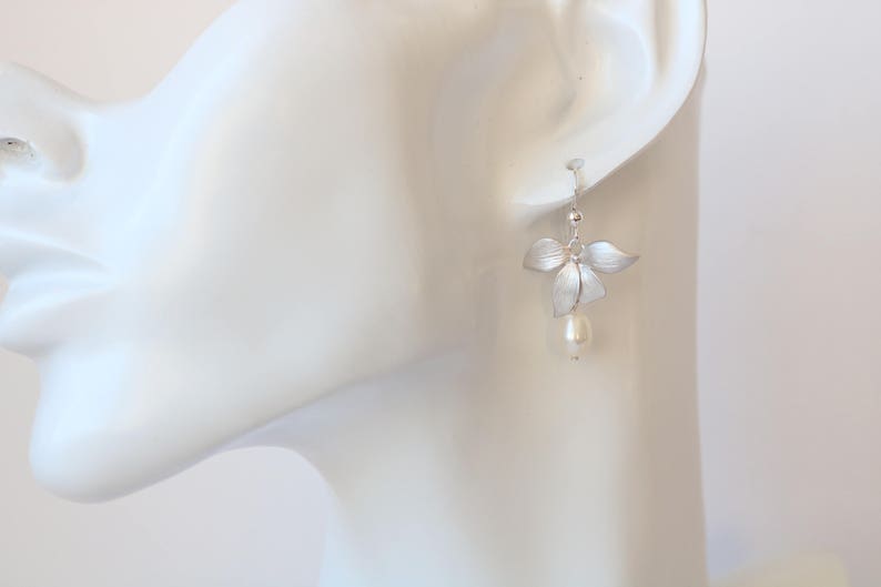 May include: Silver flower earrings with a single pearl drop. The earrings are on a white mannequin head.