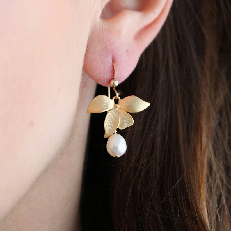 May include: Gold orchid flower earrings with a single pearl dangling below. The earrings are a delicate and feminine design.