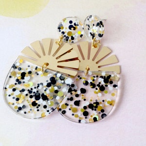 Pode incluir: Um par de brincos de acr&iacute;lico transparente e dourado com um design em forma de leque e confete preto, branco e dourado.