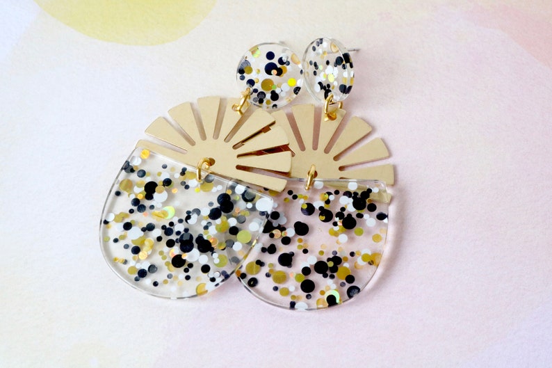 Pode incluir: Um par de brincos de acr&iacute;lico transparente e dourado com um design em forma de leque e confete preto, branco e dourado.