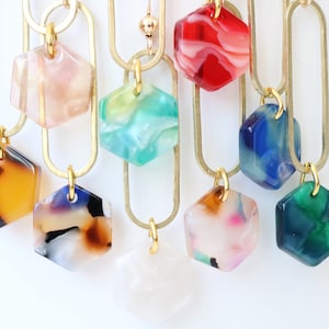 May include: A set of nine hexagon-shaped pendants with marbled patterns in various colours, including pink, green, red, blue, brown, and white. The pendants are attached to gold-coloured chains.