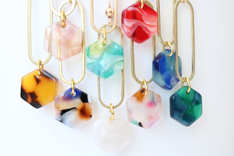 May include: A set of nine hexagon-shaped pendants with marbled patterns in various colours, including pink, green, red, blue, brown, and white. The pendants are attached to gold-coloured chains.
