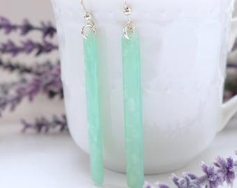 May include: A pair of long, light green dangle earrings with silver hooks. The earrings are made of a translucent material with a marbled effect.