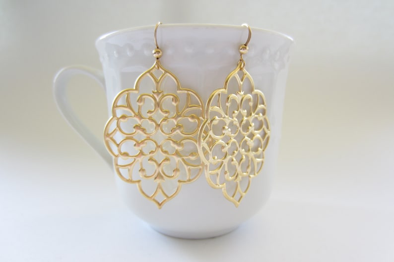 Puede incluir: Un par de pendientes de filigrana dorada con un dise&ntilde;o delicado e intrincado. Los pendientes cuelgan de una taza de cer&aacute;mica blanca.