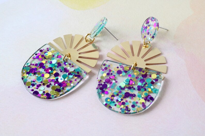 Pode incluir: Um par de brincos de resina transparente e dourada com um design de sol e confete colorido. Os brincos s&atilde;o uma pe&ccedil;a de destaque e seriam perfeitos para uma festa ou uma ocasi&atilde;o especial.