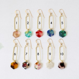 May include: Ten pairs of gold earrings with a long, oval-shaped dangle and a hexagonal charm. The charms are made of colourful, marbled resin in various colours, including red, green, blue, brown, and white.