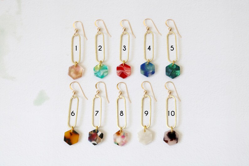 May include: Ten pairs of gold earrings with a long, oval-shaped dangle and a hexagonal charm. The charms are made of colourful, marbled resin in various colours, including red, green, blue, brown, and white.