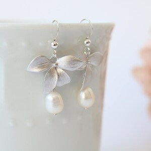 May include: Silver earrings with a delicate flower design and a pearl drop. The earrings are hanging from a white surface.