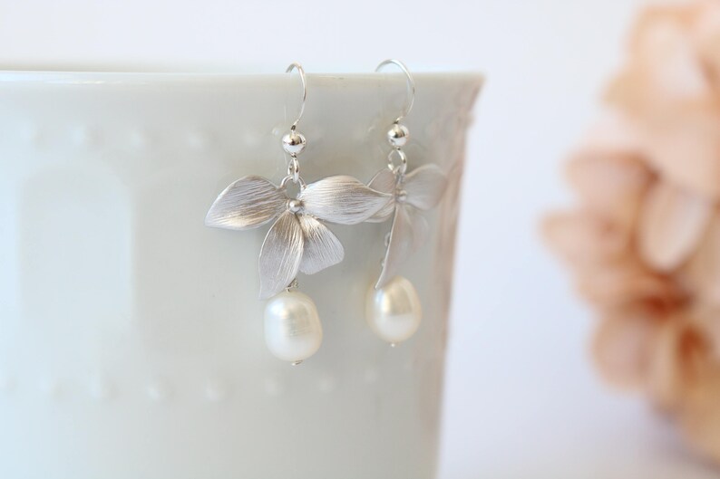 May include: Silver earrings with a delicate flower design and a pearl drop. The earrings are hanging from a white surface.