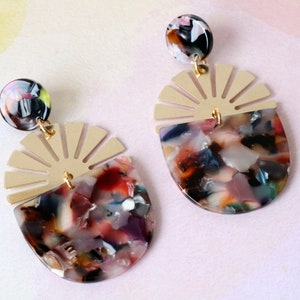 Pode incluir: Um par de brincos dourados e multicoloridos com um design de sol e um grande pingente redondo, multicolorido, marmorizado, de resina.