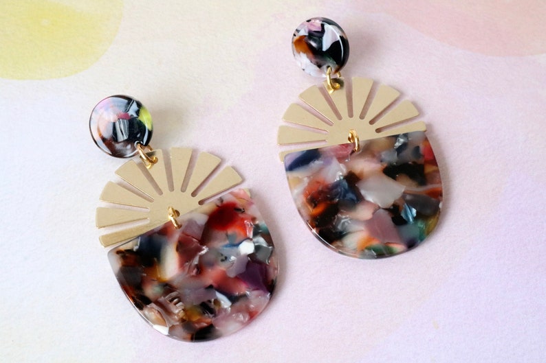 Pode incluir: Um par de brincos dourados e multicoloridos com um design de sol e um grande pingente redondo, multicolorido, marmorizado, de resina.