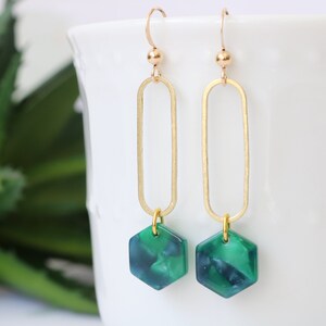 May include: Gold dangle earrings with a green hexagon-shaped pendant. The pendant has a marbled pattern.