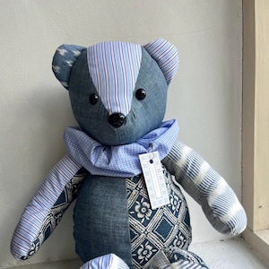 Puede incluir: Un oso de peluche azul hecho a mano con un diseño de patchwork. El oso presenta varias telas azules, incluyendo rayas y estampados florales. Tiene ojos de botón negros y un cuello azul. Una etiqueta cuelga del cuello.