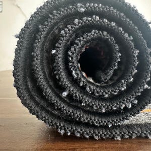 May include: Rolled-up gray rug with a black border and orange stripe. The rug is made of a textured material and is tightly rolled, revealing the layers of fabric. The image is a close-up, highlighting the rug's texture and design.