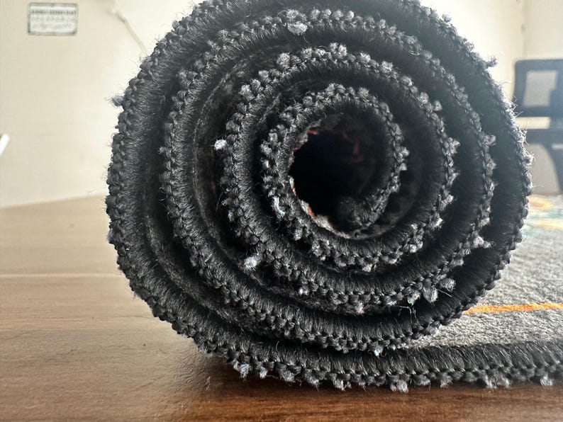 May include: Rolled-up gray rug with a black border and orange stripe. The rug is made of a textured material and is tightly rolled, revealing the layers of fabric. The image is a close-up, highlighting the rug's texture and design.