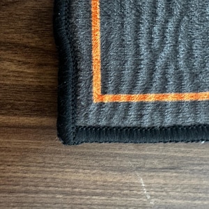 May include: Close-up of a dark gray textile with an orange border and black stitching. The fabric has a textured, ribbed appearance. The orange border creates a contrasting accent against the darker gray material.
