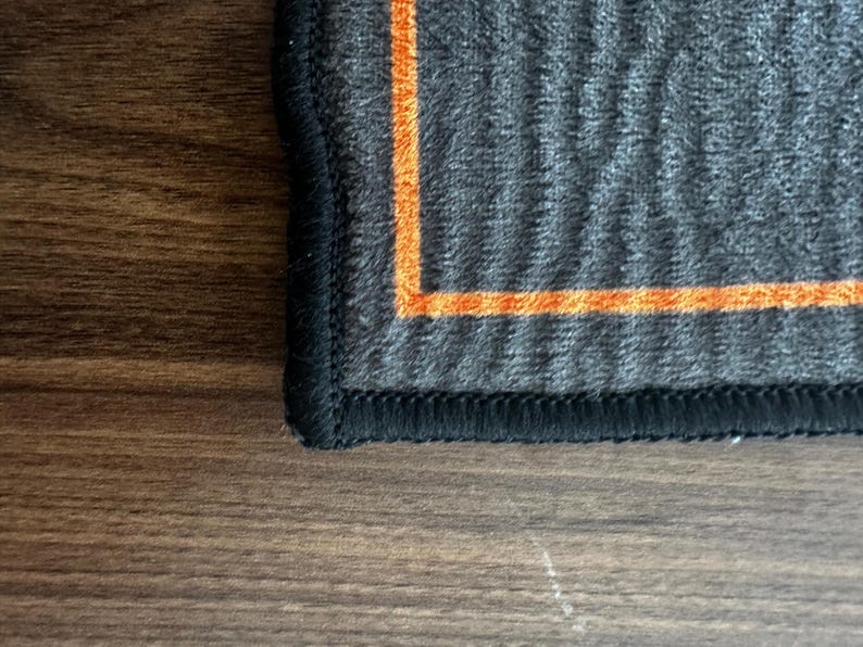May include: Close-up of a dark gray textile with an orange border and black stitching. The fabric has a textured, ribbed appearance. The orange border creates a contrasting accent against the darker gray material.