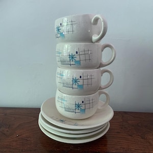 May include: A stack of four off-white ceramic cups with handles and four matching saucers. Each piece features a speckled finish and a geometric pattern consisting of thin black grid lines, grey rectangular blocks, and blue starburst accents.