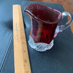 Könnte beinhalten: Ein kleiner, rubinroter Glaskrug mit einem Griff und einer Basis aus klarem Glas. Der Krug hat einen strukturierten Boden im Hobnail-Stil und einen ausgestellten Ausguss. Ein Holzlineal liegt neben dem Krug.