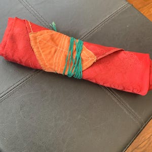 Op de afbeelding: Een opgerold rood stoffen item met een oranje blad-vormige patch. Het item is vastgezet met groen touw. De rode stof heeft een subtiel patroon. De oranje patch is gestikt met wit garen.