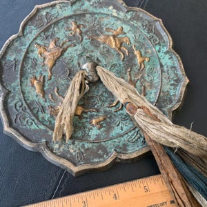 Op de afbeelding: Antieke decoratieve plaat van bronskleurig metaal met een geschulpte rand en een groen patina. De plaat heeft verhoogde reliëf figuren van mensen te paard en andere dieren. Twee kwasten met lederen en blauwe accenten zijn bevestigd.