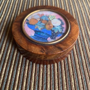 May include: A round wooden box with a polished brown grain finish. The lid features a circular inset with a colorful painted illustration of various fruits, including oranges and grapes, arranged in a blue bowl against a soft pink background.