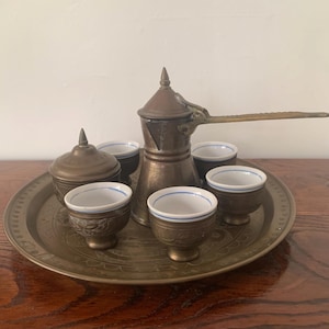 Peut inclure: Un service à café en laiton vintage, comprenant une cafetière, de petites tasses et un récipient avec couvercle, disposés sur un plateau rond. Les tasses ont des intérieurs blancs avec des bords bleus. L'ensemble est probablement pour le café turc ou arabe.