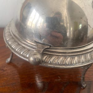 Può includere: Un burriera antica color argento con coperchio a cupola e dettagli decorativi in rilievo. Il piatto ha una base rotonda con un bordo decorativo e piccoli piedini curvi. Una piccola maniglia è attaccata al lato.