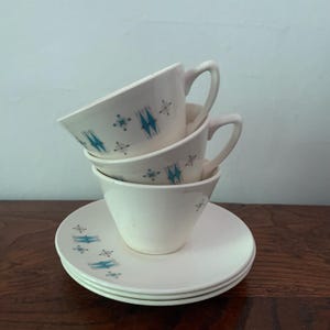 May include: A stack of two off-white ceramic teacups with handles, resting on a pair of matching saucers. The cups and saucers feature a repeating pattern of small teal diamond shapes and thin gray starburst lines across the cream-colored surface.