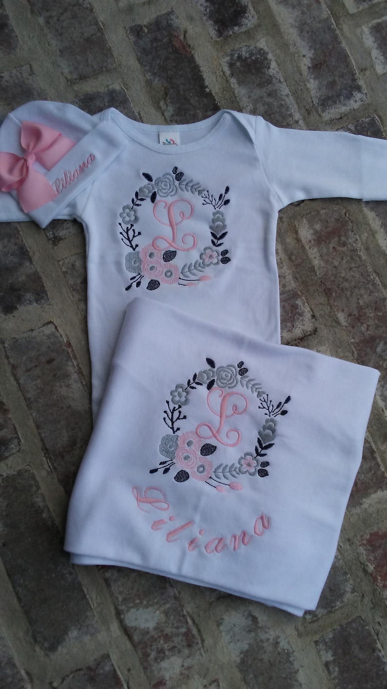 May include: A white baby outfit with a pink and gray floral wreath embroidered with the letter "L" and the name "Liliana". The outfit includes a long-sleeved onesie and a blanket.