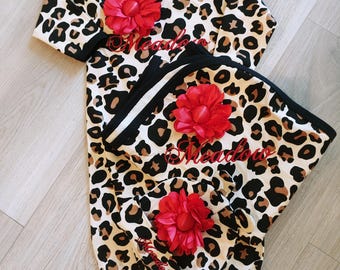 May include: A leopard print baby gown and matching hat set with a red flower embellishment and the name "Meadow" embroidered in red on each piece.